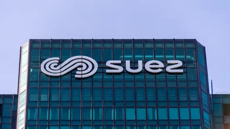 The Suez corporate building showing its white branded logo and name on a teal glass facade against a light sky